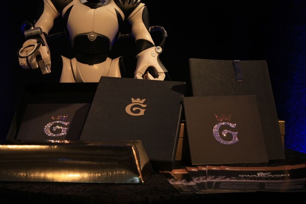 Ein Tisch vor blauem Bühnenlicht: dunkle Präsentationsmappen mit goldenem Monogramm, dahinter eine Roboterfigur.